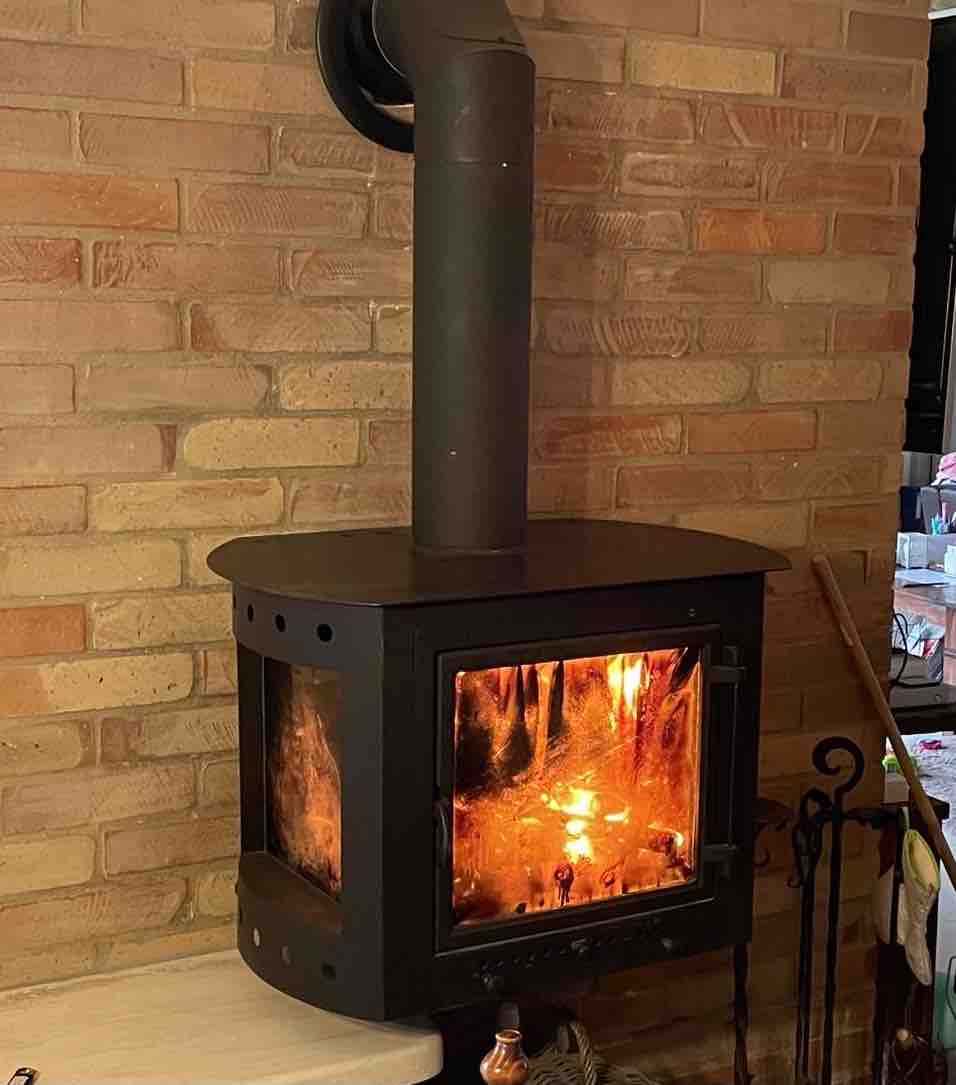 A sleek wood-burning stove is mounted on a brick wall, featuring a glass front showcasing a warm glow from the flames inside. The simple design adds an element of comfort to the surrounding space.