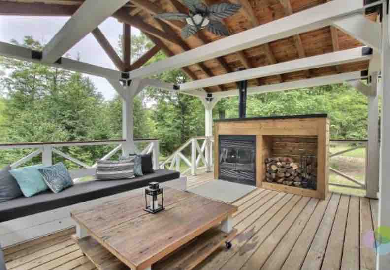 An outdoor terrace is showcased, featuring a comfortable seating area with a dark sofa adorned with decorative cushions. A wooden coffee table sits at the center, and a stone fireplace is built into one side, with stacked logs nearby. Lush greenery surrounds the space.
