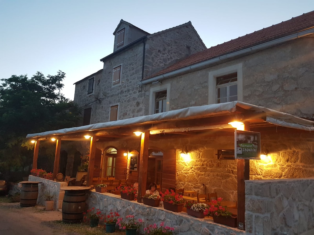 A rustic stone building features a welcoming tavern entrance adorned with warm lighting. Potted flowers line the wooden terrace, enhancing the charm of the outdoor seating area. The structure exhibits a historical character, complemented by wooden barrels placed beside the entry.