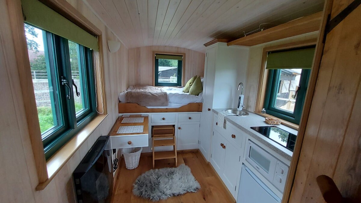 The interior of the wagon features wooden paneling and large windows, allowing natural light to fill the space. A compact kitchenette is equipped with a microwave and hob, while a cozy sleeping area is set with a small double bed beside a small table and chair.