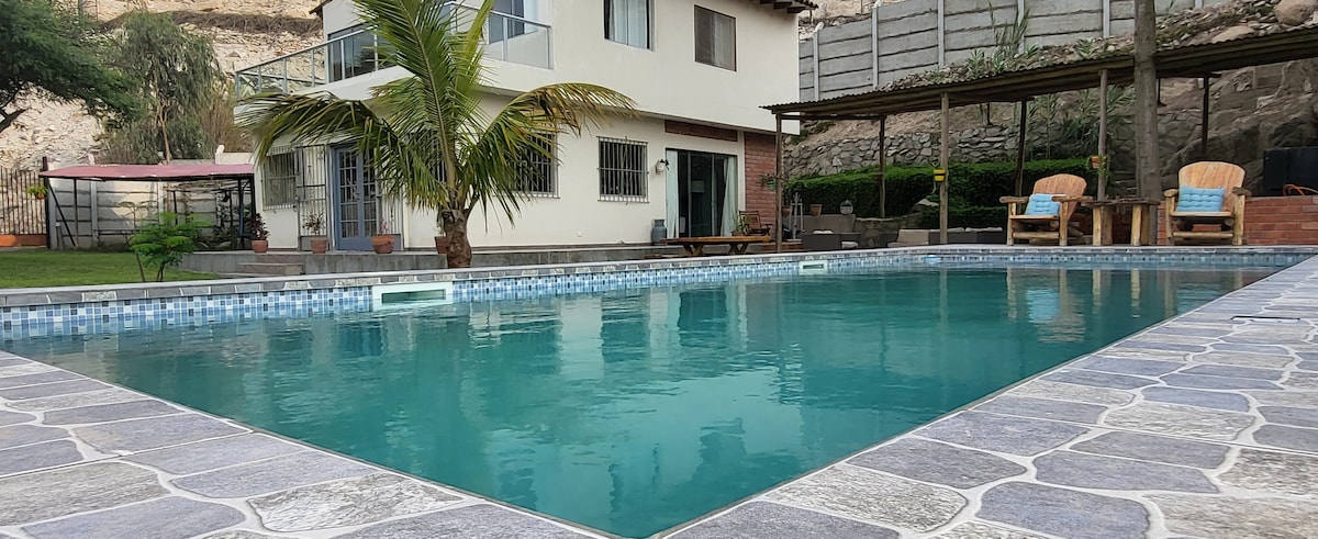 A spacious outdoor swimming pool is framed by a terrace featuring seating areas. Lush greenery, including a palm tree, adds a natural touch. The house with large windows is visible in the background, providing a welcoming view.