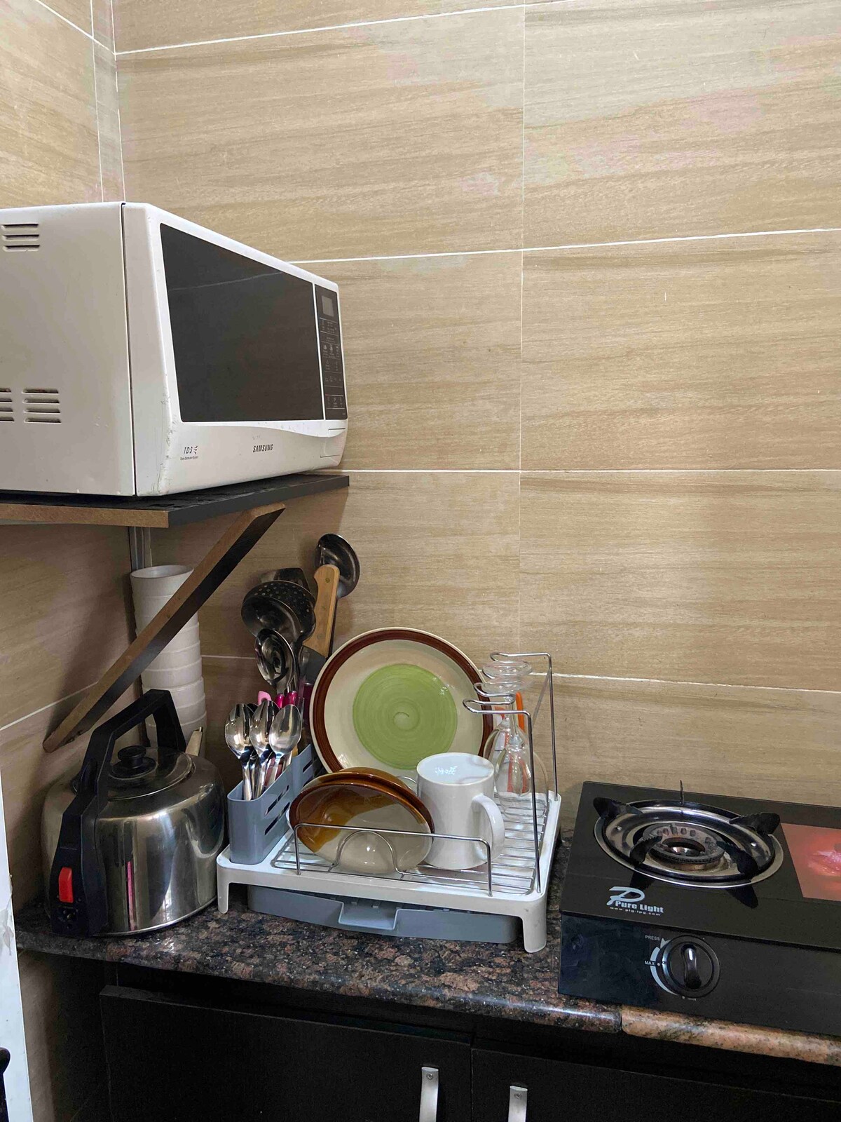 A cozy kitchen corner features a countertop with various utensils, including knives and spoons. The neatly arranged dishes, including plates and glasses, sit on a drying rack beside a compact gas stove and a microwave above, creating a functional cooking space.