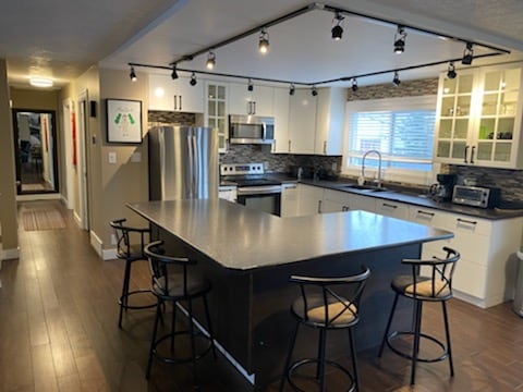 A modern kitchen is displayed with sleek cabinetry and stainless steel appliances. An island with bar stools provides casual seating. Track lighting highlights the space, while a window allows natural light to brighten the room. The layout offers a seamless flow to adjacent areas.