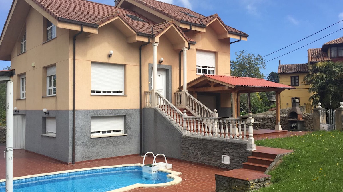 A spacious villa is shown with a private pool and a well-maintained garden. Stairs lead to a covered terrace area, with a barbecue station visible nearby. The exterior features a combination of light and dark colors, and large windows allow for ample natural light.