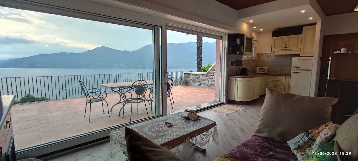 A spacious living area features large glass doors that open to an outdoor terrace with round dining tables and chairs. Scenic views of the mountains and lake are visible through the doors. Soft furnishings add comfort to the interior space.