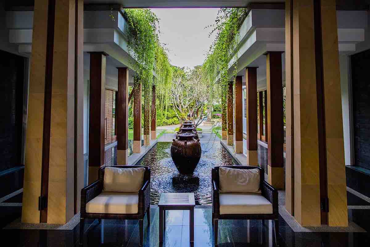 An entrance area features two comfortable seating arrangements with soft cushions, facing a tranquil water feature. Lush greenery is visible in the background, framed by elegantly designed columns that enhance the open atmosphere.