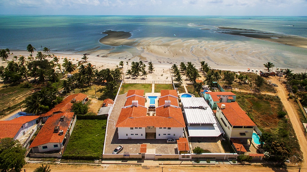 An aerial view captures the expansive coastline with gentle waves lapping at the sandy shore. The property features a layout of several buildings with red-tiled roofs surrounding a central pool. Lush greenery and palm trees enhance the tropical ambiance.