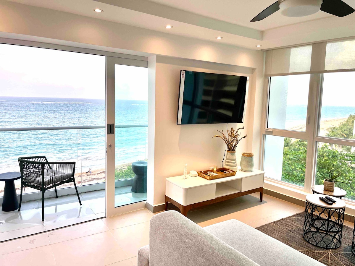 A well-lit living area features a large flat-screen TV mounted on the wall and a stylish console beneath. Full-length windows offer a direct view of the Atlantic Ocean, while a comfortable seating area is arranged beside a small balcony with outdoor furniture.