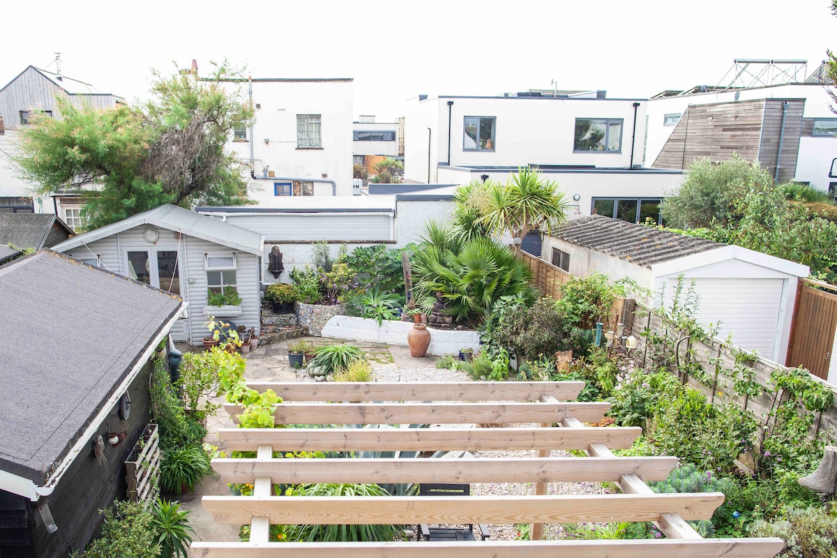 A well-maintained garden area offers a variety of plants and features, including a patio space lined with wooden trellises. A small shed is positioned in one corner, with neighboring modern buildings visible in the background. Multiple greenery elements create a relaxing outdoor environment.