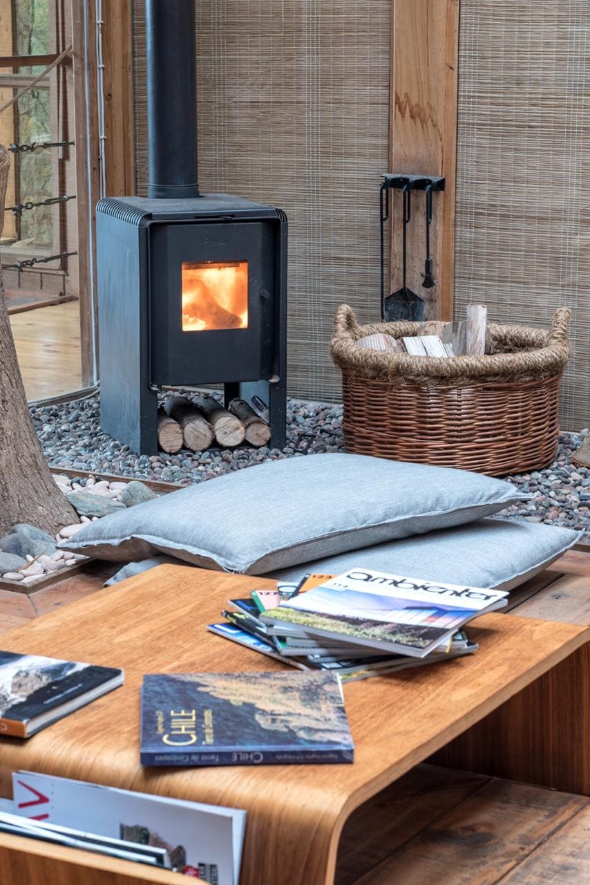 A cozy living area features a wood-burning stove with a warm glow, surrounded by a woven basket holding firewood. A coffee table is adorned with several magazines, and a plush cushion is placed nearby for comfortable seating. Natural materials and textures are highlighted throughout.
