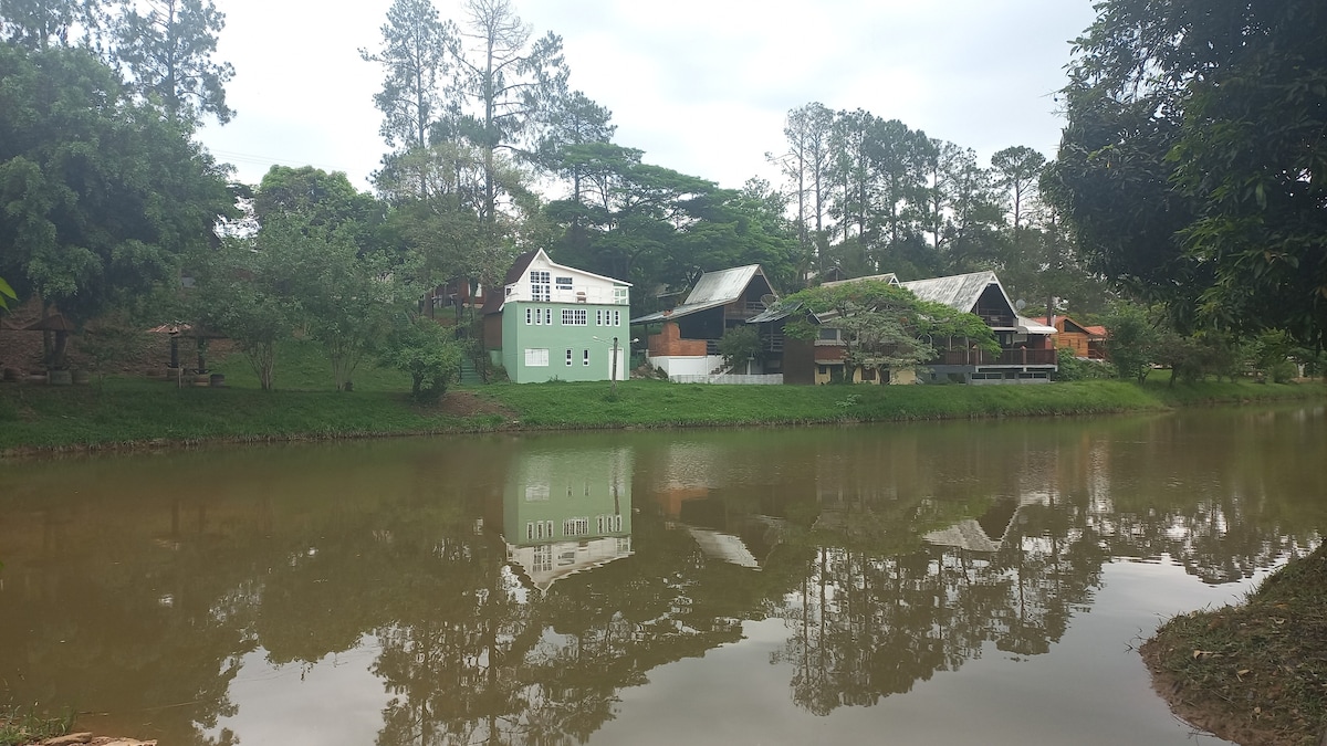A serene lakeside view showcases a group of rustic structures nestled among trees. The water reflects the green and wooden buildings, creating a calm ambiance. Natural surroundings enhance the peaceful setting, offering a sense of tranquility and connection to nature.