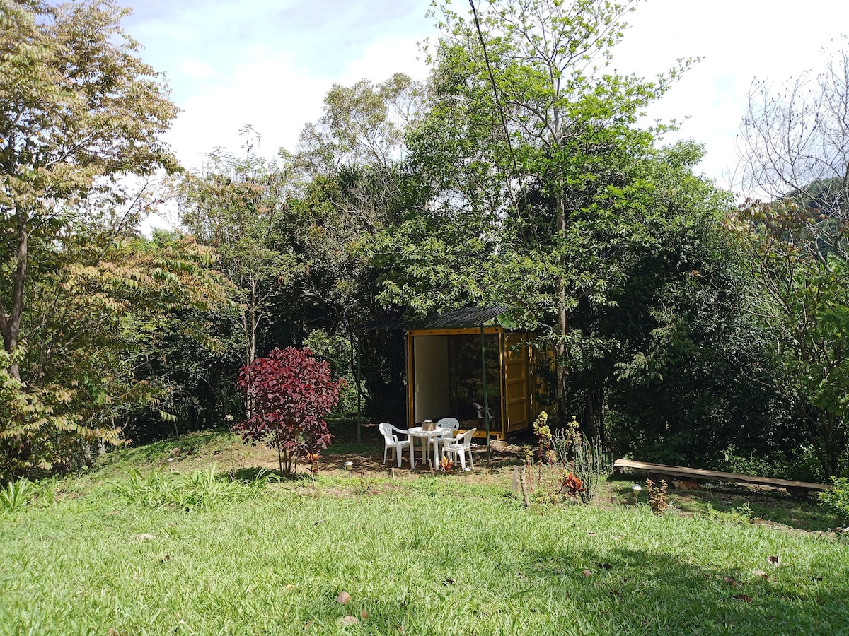 A small, bright structure is nestled in a lush green landscape, surrounded by trees. A seating area with white chairs is set outside, providing an inviting space to enjoy the natural surroundings. Vibrant foliage adds color to the serene environment.