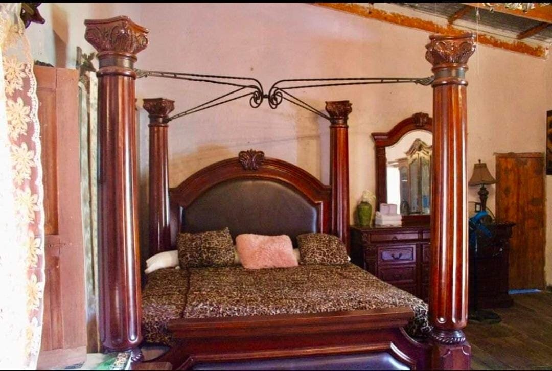 A spacious bed with an ornate wooden frame featuring four decorative posts is highlighted. Soft bedding in warm tones is layered across the bed. A wooden dresser and a mirror are positioned nearby, adding to the room's classic design.