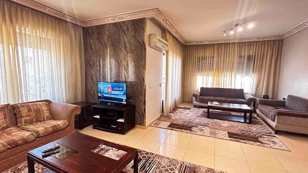 A spacious living area is presented, featuring large windows with light drapes that allow natural light to fill the room. Soft couches and a wooden coffee table are arranged for comfort, while a television is positioned on a dark media unit. Elegant wall decor adds to the ambiance.