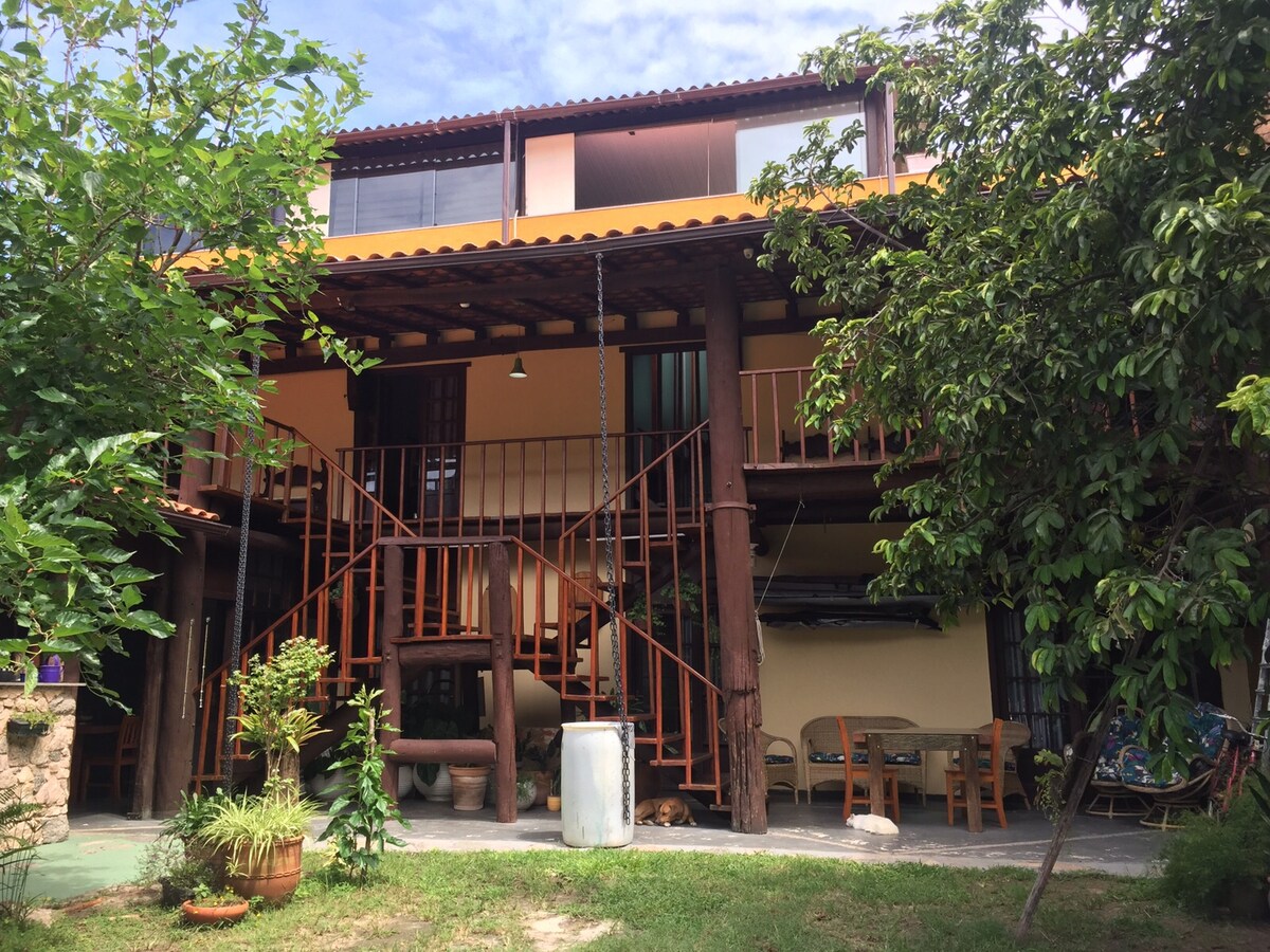 The exterior of a rustic three-story house is displayed, featuring wooden beams and balconies. Surrounding greenery includes trees and plants. A staircase leads to the upper levels. An outdoor seating area with tables is visible, enhancing the welcoming ambiance of the space.