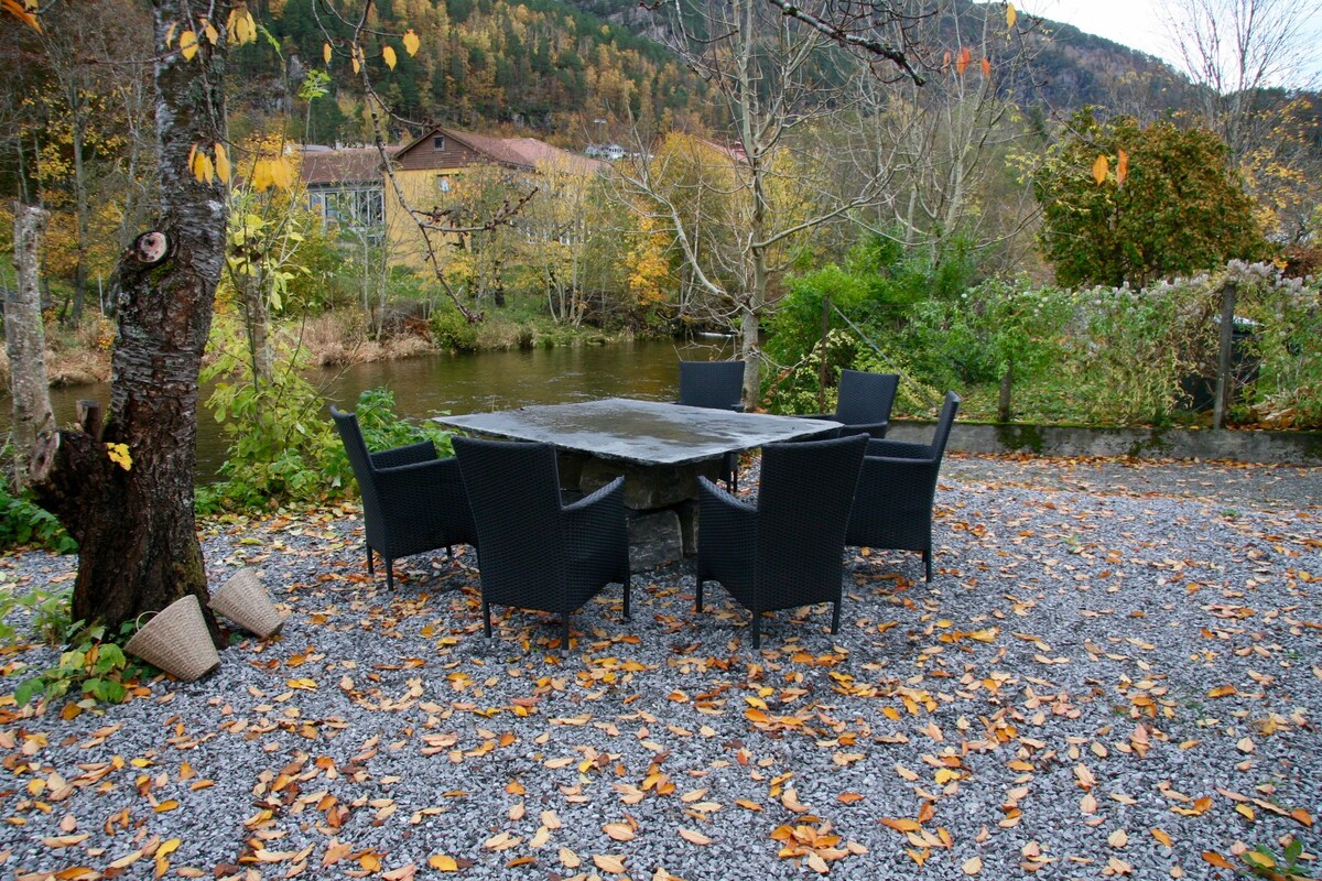 An outdoor seating area features a rectangular stone table surrounded by six dark wicker chairs. The table is situated on a gravel surface, with fallen autumn leaves scattered around. A view of a calm river and greenery can be seen in the background.