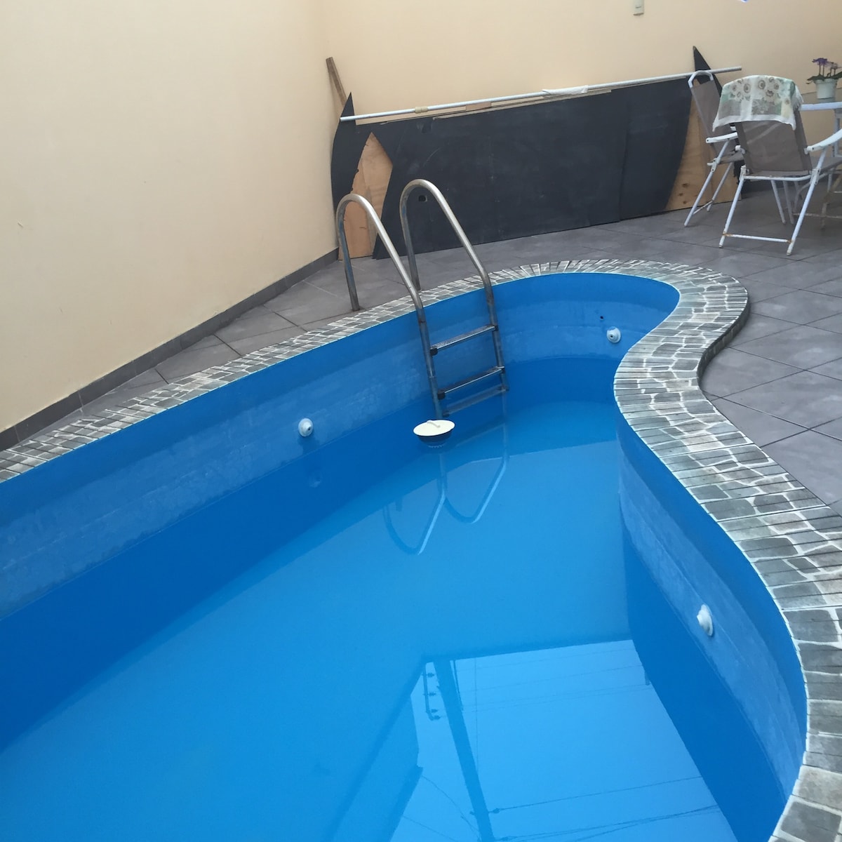 A private swimming pool with a curved design is set against a neutral-colored wall. Steps lead into the clear blue water, and surrounding tiles enhance the space. A small table and chairs are positioned nearby, offering a spot for relaxation.