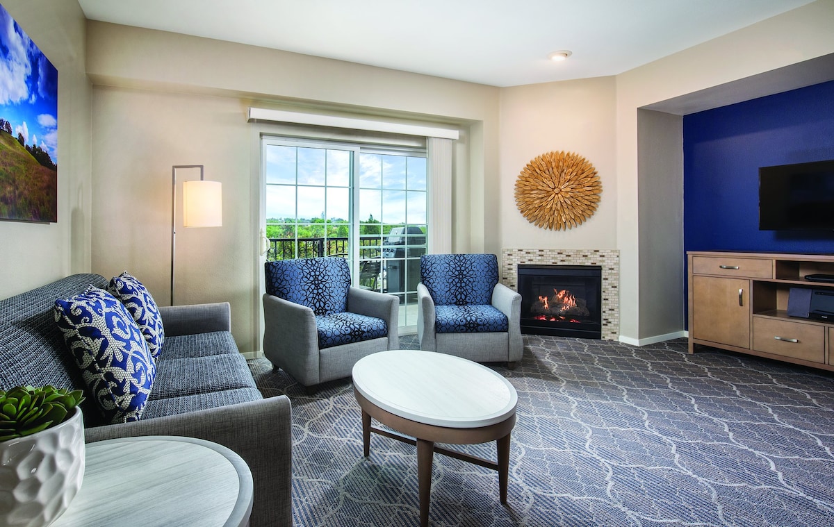 A comfortable living area is presented with a gas fireplace, a round coffee table, and two armchairs alongside a sofa. Natural light fills the space through large windows, which offer a view of the outdoors. A flat-screen TV is mounted on the wall across from the seating.