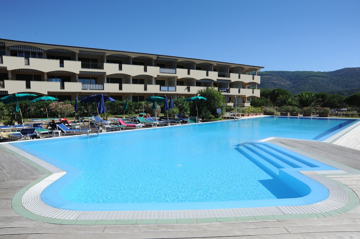 A wide, clear swimming pool is featured in the foreground, surrounded by lounge chairs and umbrellas. The multi-story building is visible in the background, set against a clear blue sky and green hills, creating a peaceful atmosphere.