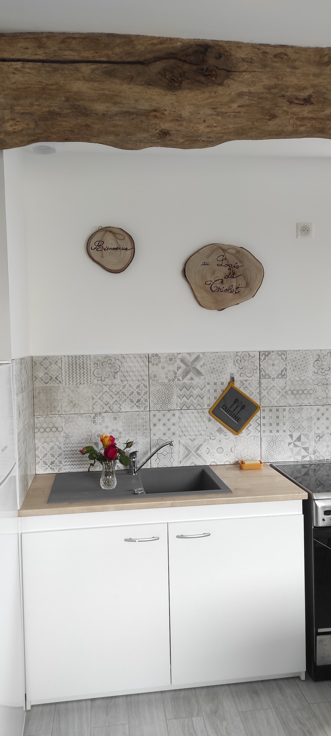 The kitchen features a modern design with clean white cabinets and a light wood countertop. Natural wood accents decorate the wall above the sink, complemented by decorative tiles with intricate patterns. A small bouquet of flowers is placed on the counter, adding a touch of color.