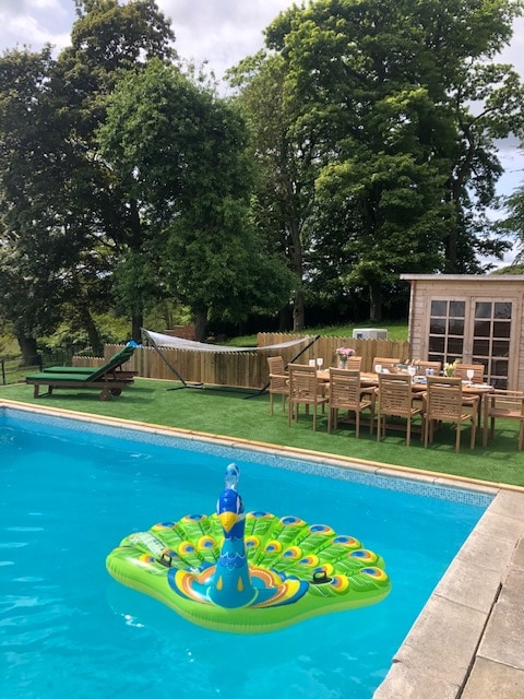A swimming pool is surrounded by lush greenery, featuring a large inflatable peacock float. Nearby, a table is set for outdoor dining, with wooden chairs arranged to accommodate guests. A garden shed is visible in the background, enhancing the inviting outdoor space.