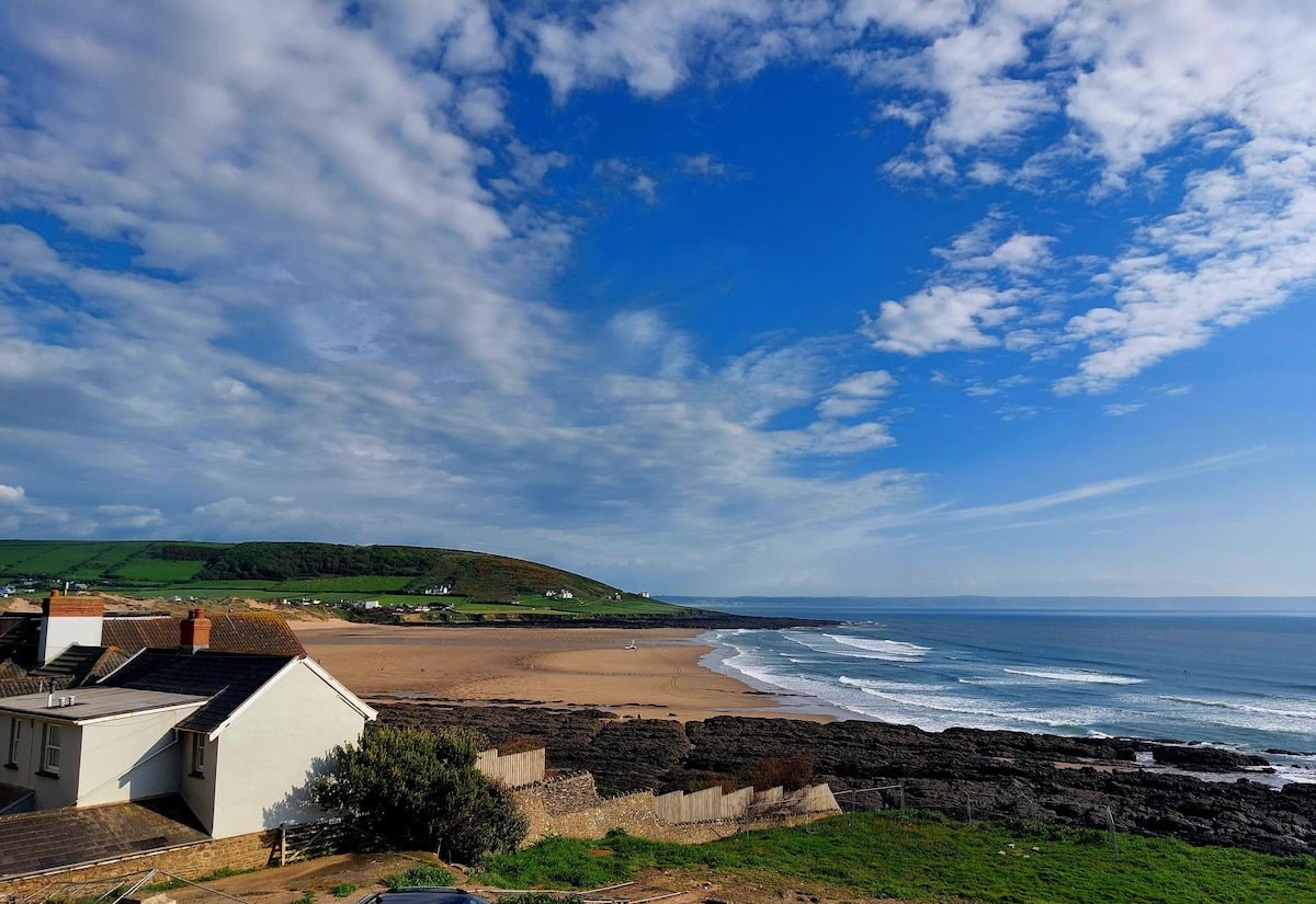 A panoramic view captures the expansive Croyde Bay with its sandy beach and gentle waves. Green hills rise in the distance under a partly cloudy sky. The scene features a part of a building in the foreground, complementing the natural landscape.