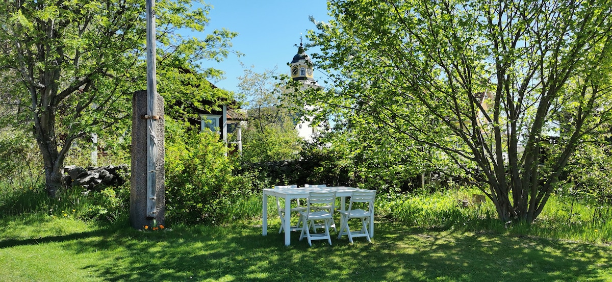 A serene outdoor seating area is set up beneath leafy trees, featuring a white table accompanied by four chairs. The surroundings exhibit a lush green lawn and a glimpse of a rustic building in the background, adding to the peaceful ambiance.