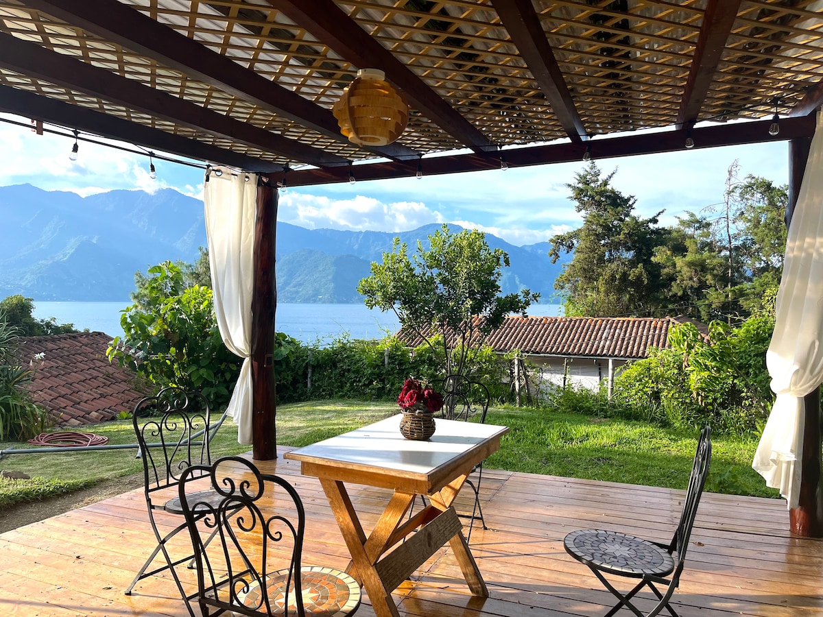 An outdoor dining area features a wooden table surrounded by four black metal chairs. A view of the lake and mountains is visible in the background, framed by greenery. Soft curtains are draped around the structure, enhancing the space's comfortable ambiance.