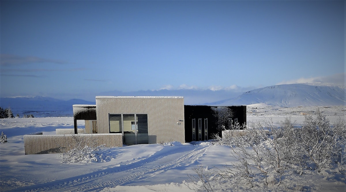 A modern house is depicted in a snowy landscape, featuring a sleek exterior with wooden paneling. The structure includes multiple windows and a covered entrance, surrounded by untouched snow and gentle slopes in the background.