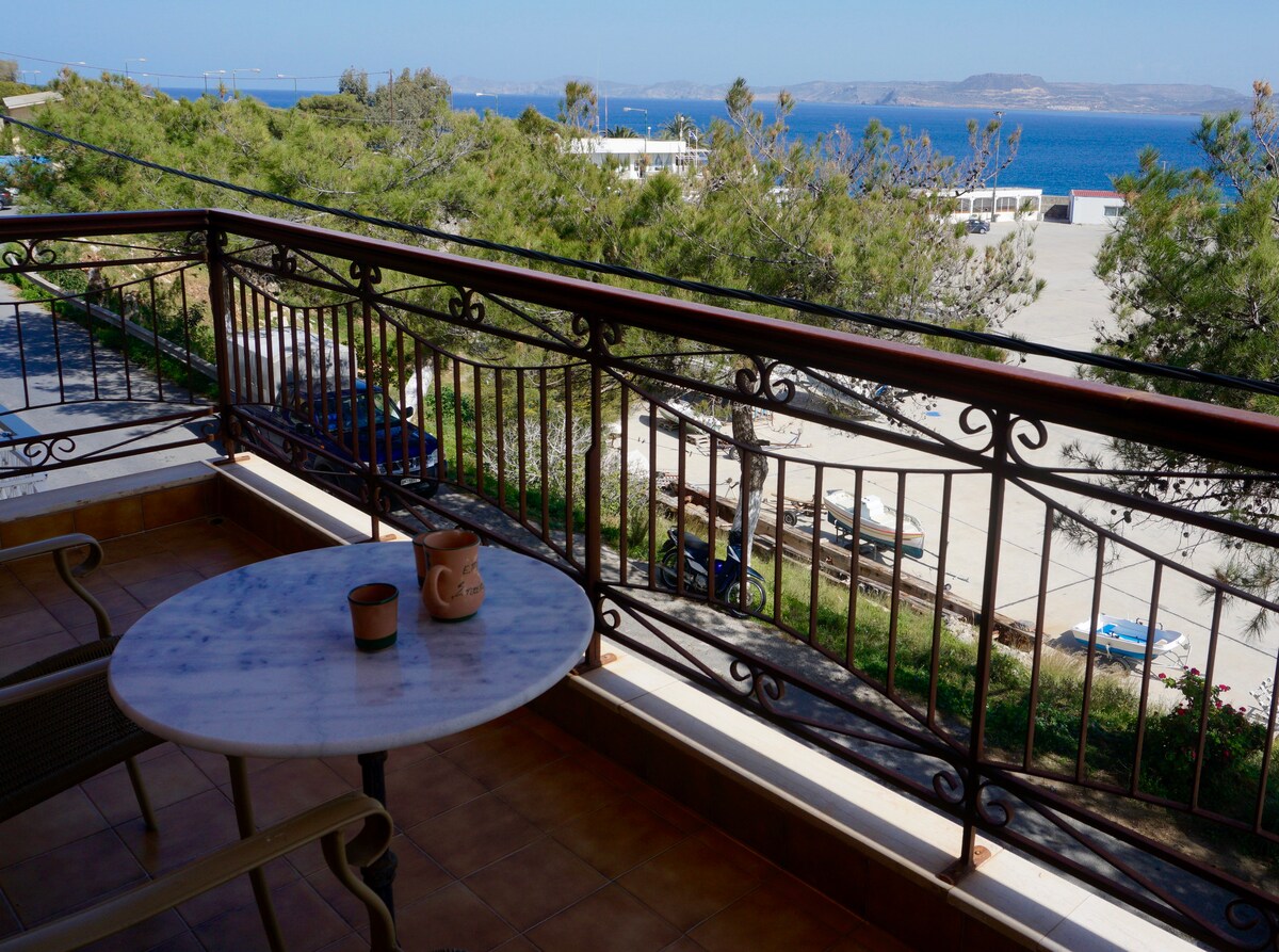 A balcony features a small marble table and a potted plant, overlooking a scenic sea view. The railing provides a clear sightline to the water and distant hills, enhancing the overall ambiance of the outdoor space.