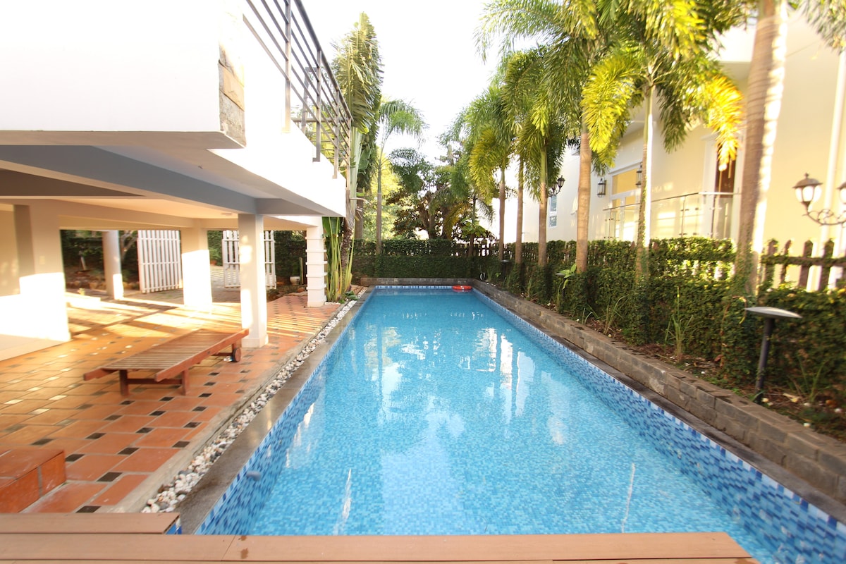 A serene swimming pool is positioned adjacent to a charming terrace. Lush palm trees surround the area, providing shade and enhancing the tranquil setting. The water reflects the clear sky, while the terracotta tiles and wooden benches create a welcoming outdoor space.