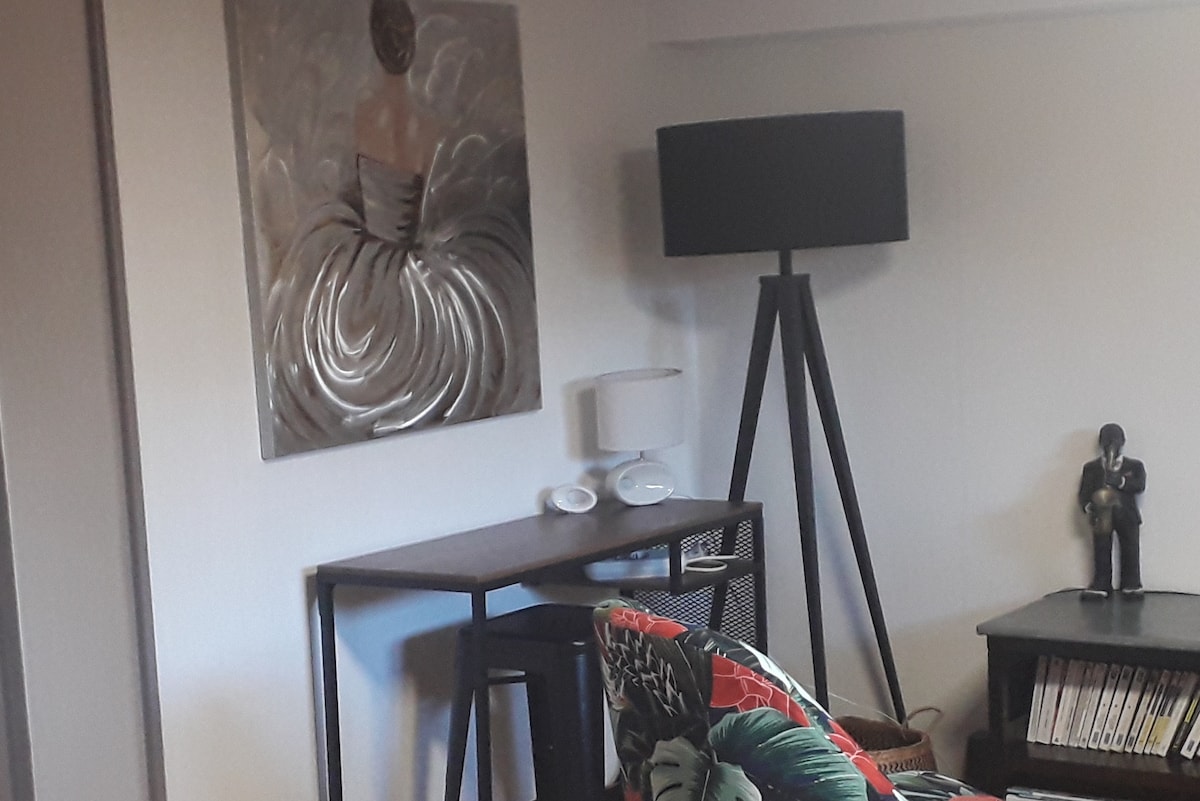 A corner of the studio features a stylish artwork depicting a dressed figure, complemented by a modern lamp. A slender table is positioned against the wall, while a decorative armchair with vibrant foliage patterns adds a pop of color to the space.