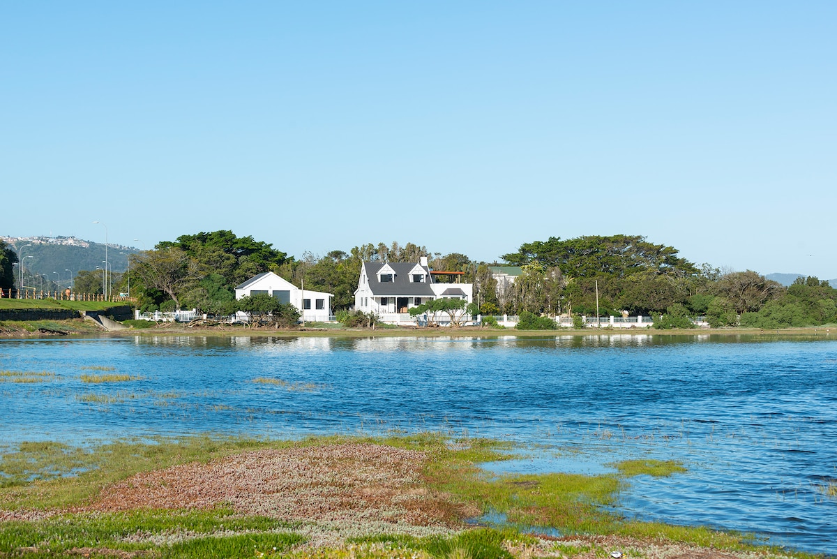 A serene view showcases a modern house situated along the Knysna Lagoon. Lush greenery surrounds the property, with gentle waves lapping at the shore. Clear blue skies reflect on the water, creating a peaceful atmosphere that invites relaxation.