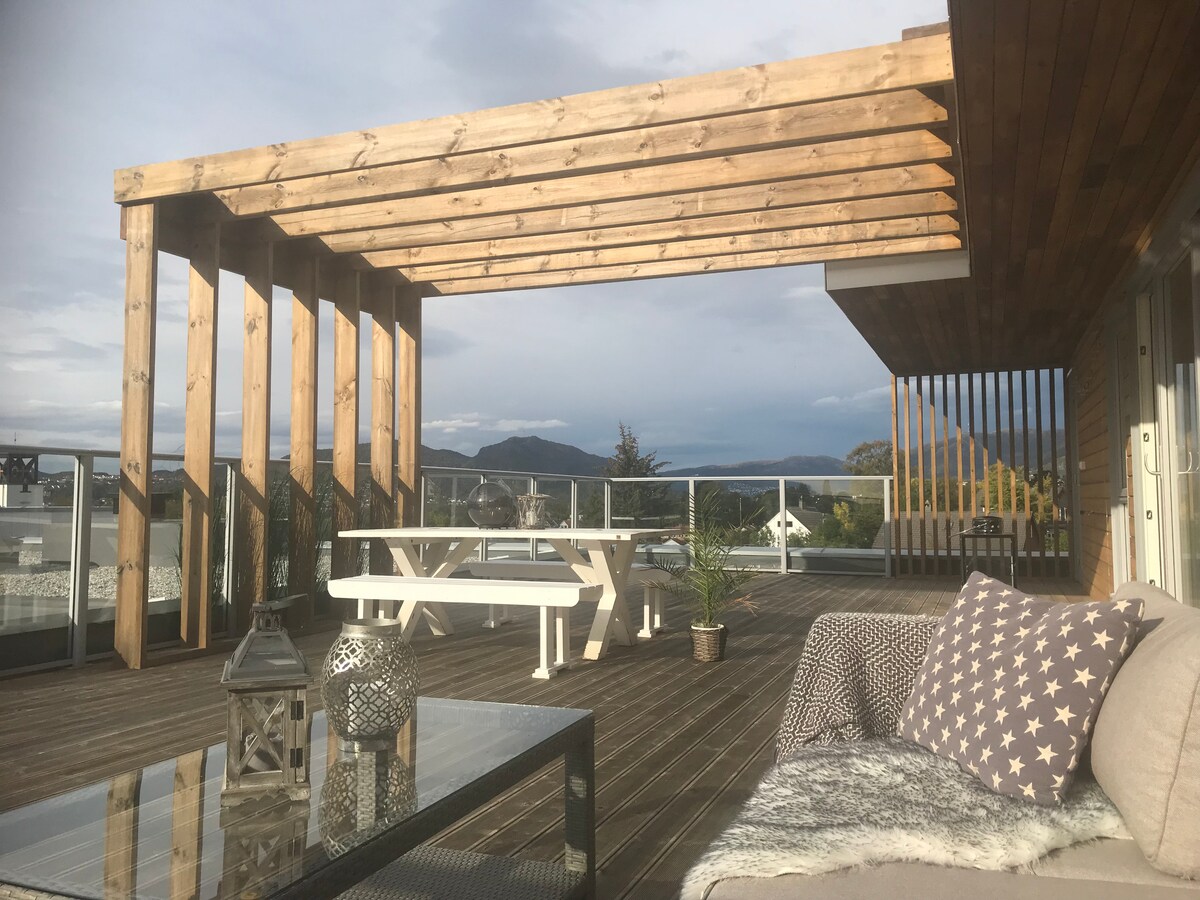 A spacious private terrace is presented, featuring a wooden pergola, outdoor seating, and a dining table. The area is surrounded by glass railings, offering unobstructed views of the distant mountains and landscape. Warm sunlight illuminates the deck, highlighting its inviting and open atmosphere.