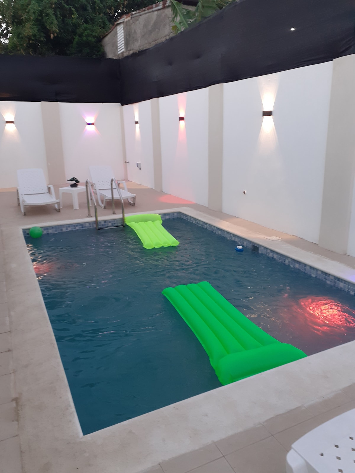 A private pool area features vibrant green floaties positioned on the water. Two white lounge chairs are placed nearby, with wall sconces providing soft lighting. The setting is enclosed by sleek walls, creating a serene atmosphere for relaxation.