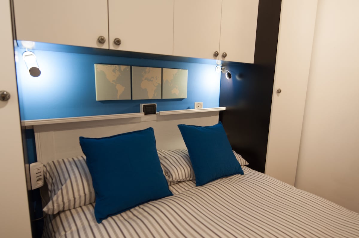 A compact sleeping area is highlighted by a comfortable bed adorned with striped linens and two blue accent pillows. A soft glow emanates from mounted lights above, illuminating the wall decorated with a world map. Ample storage is provided in the surrounding cabinetry.