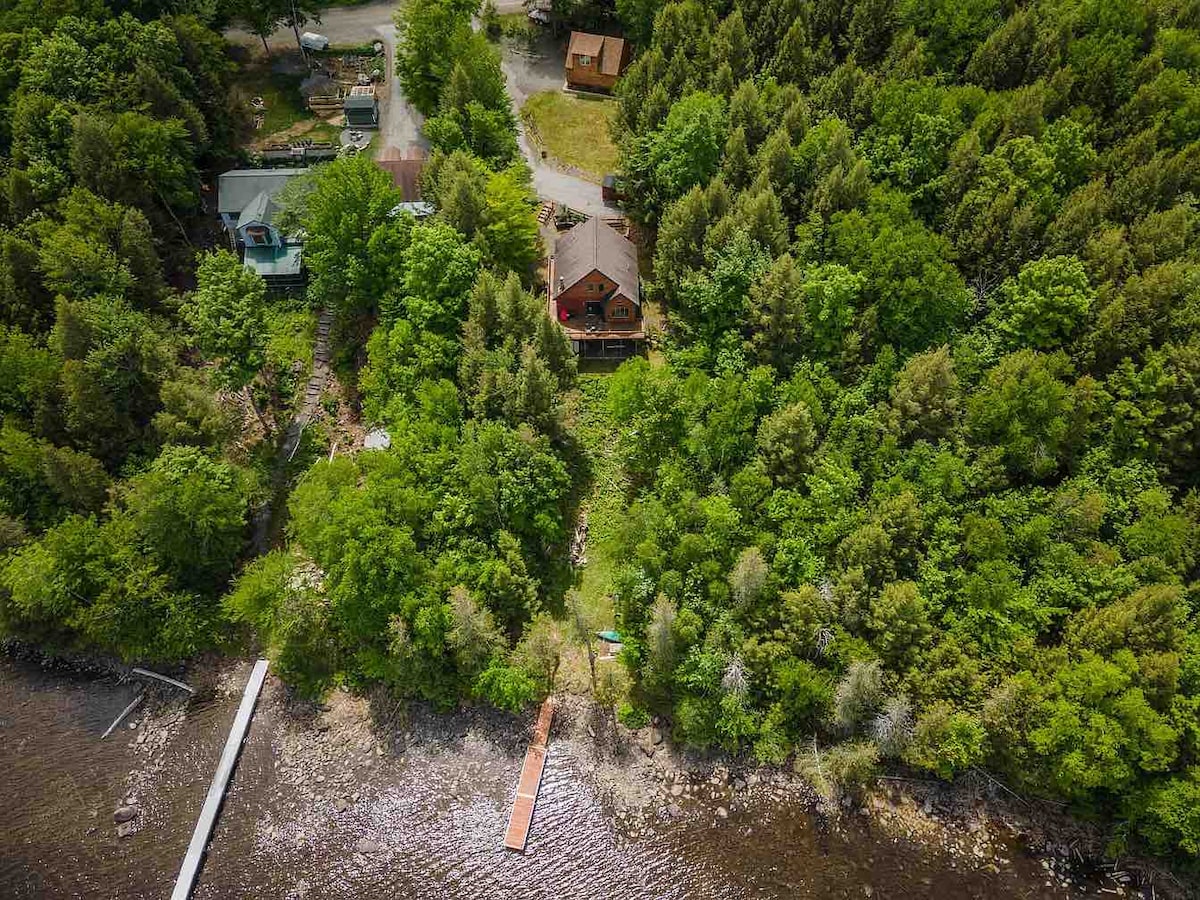 An aerial view captures a wooded landscape leading to Sebec Lake. A private dock extends into the clear water, while a cozy home is nestled among lush greenery. Nearby roads and other rustic homes are visible along the tranquil shore.