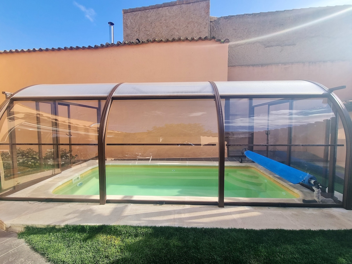 A private pool is housed under a protective enclosure, featuring clear panels that provide an unobstructed view of the serene water. Lush greenery surrounds the pool area, enhancing the inviting atmosphere for relaxation. A blue pool cover is neatly rolled up beside the pool.