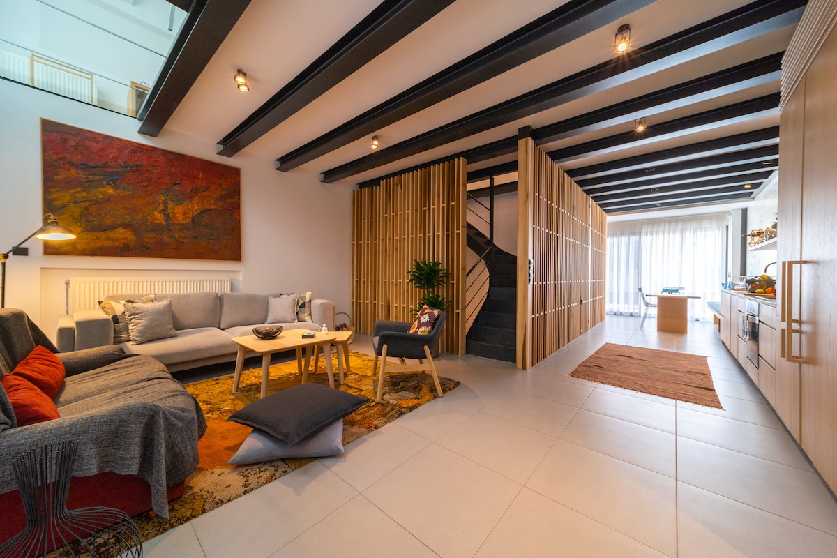 A modern living space features a comfortable seating area with a gray sofa and colorful cushions. A wooden coffee table is positioned on a patterned rug. Natural light floods in through large windows, illuminating the minimalistic kitchen and staircase that leads to the upper levels.