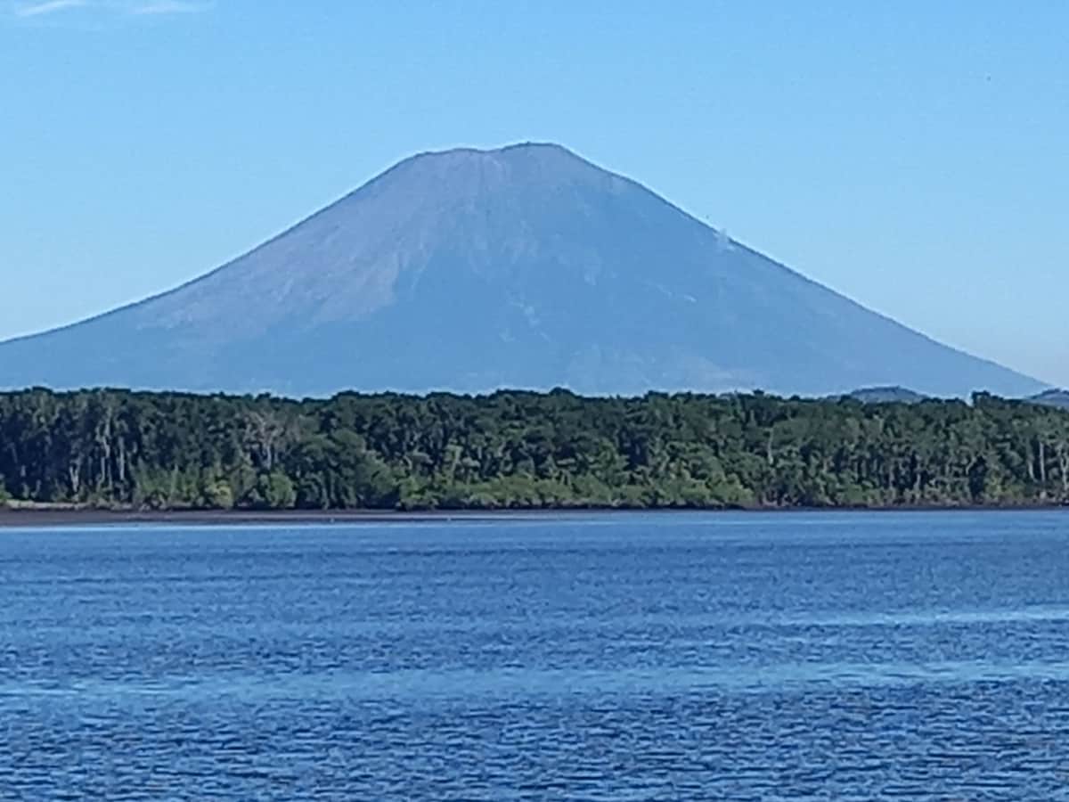 A serene scene is depicted, with a prominent volcano rising in the distance against a clear blue sky. A calm body of water reflects the lush greenery at the base of the volcano, creating a tranquil atmosphere.