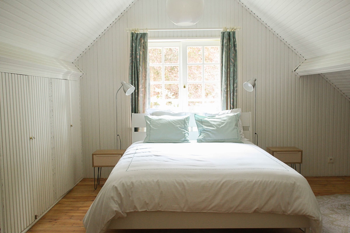 A spacious sleeping area features a large bed with soft, light-colored linens. Two nightstands are positioned on either side of the bed, with reading lamps providing illumination. Natural light fills the room through a window adorned with light curtains, while the sloped ceiling adds character.