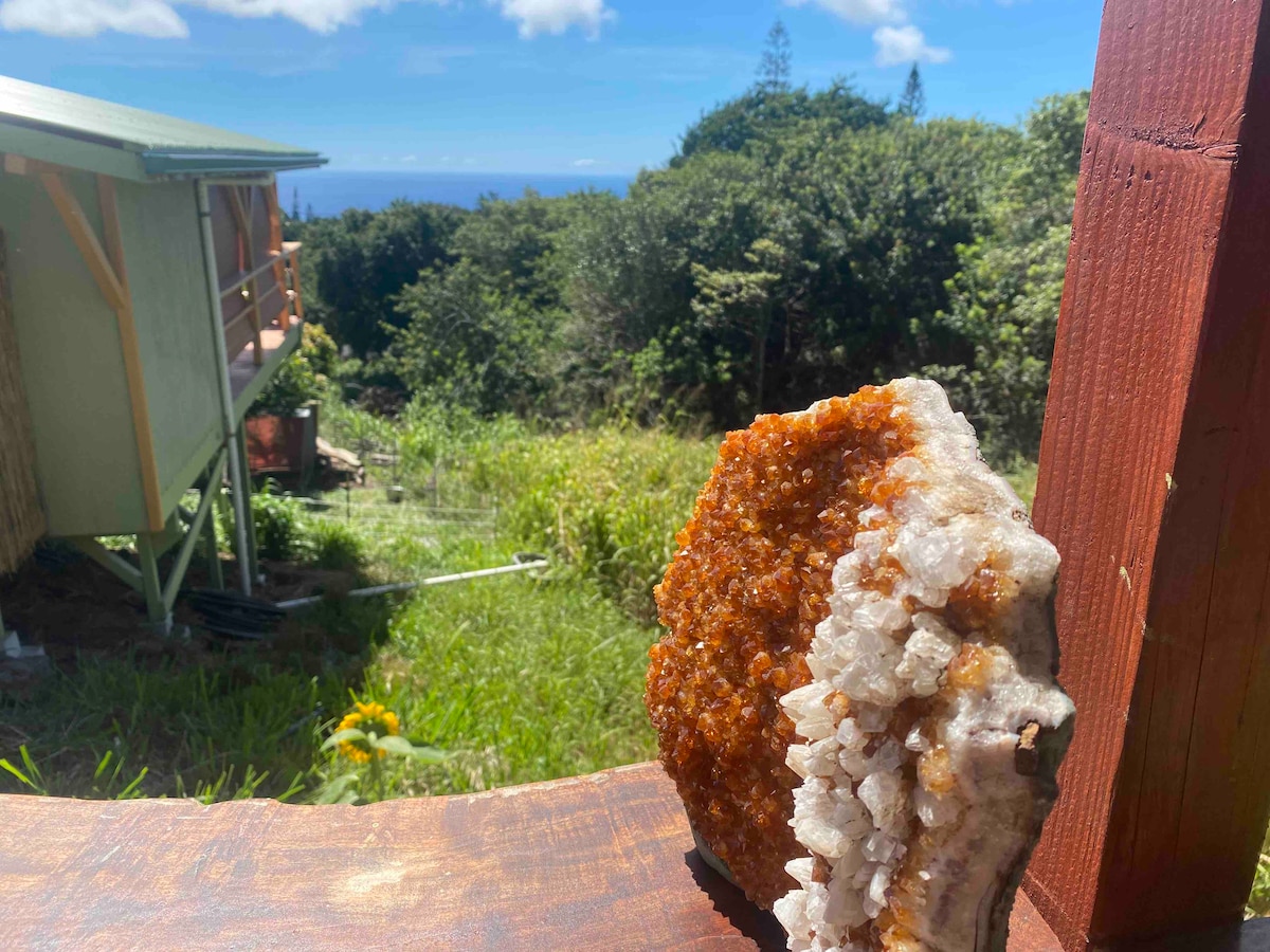 A large crystal geode sits on a rustic wooden surface, showcasing its rich amber and clear quartz formations. In the background, the cabin is partially visible, framed by vibrant greenery and a glimpse of the ocean under a bright blue sky.