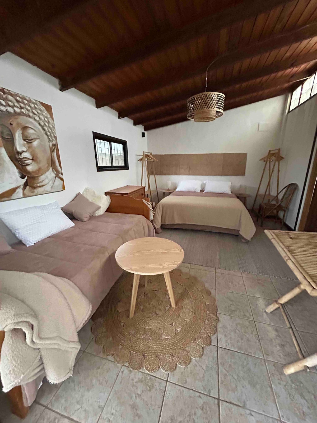 An inviting studio features a spacious arrangement with a double bed and a single bed that doubles as a sofa. Natural light filters through the windows, illuminating warm wood accents and textured decor. A small round table and woven rug enhance the cozy atmosphere.