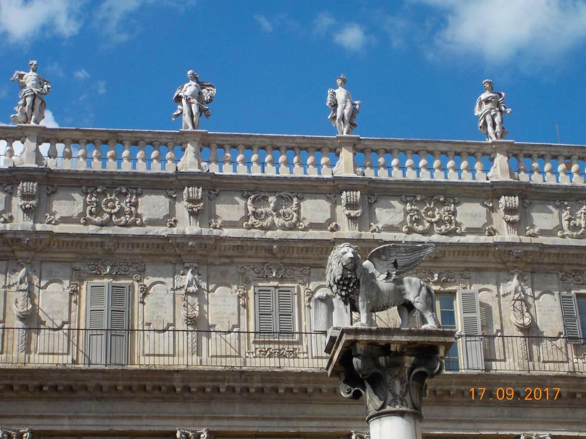 A historical building features intricate architectural details, showcasing sculpted figures on the balcony. A statue of a lion stands prominently on a column in the foreground, while windows with shutters are visible above the ornate façade under a blue sky with scattered clouds.