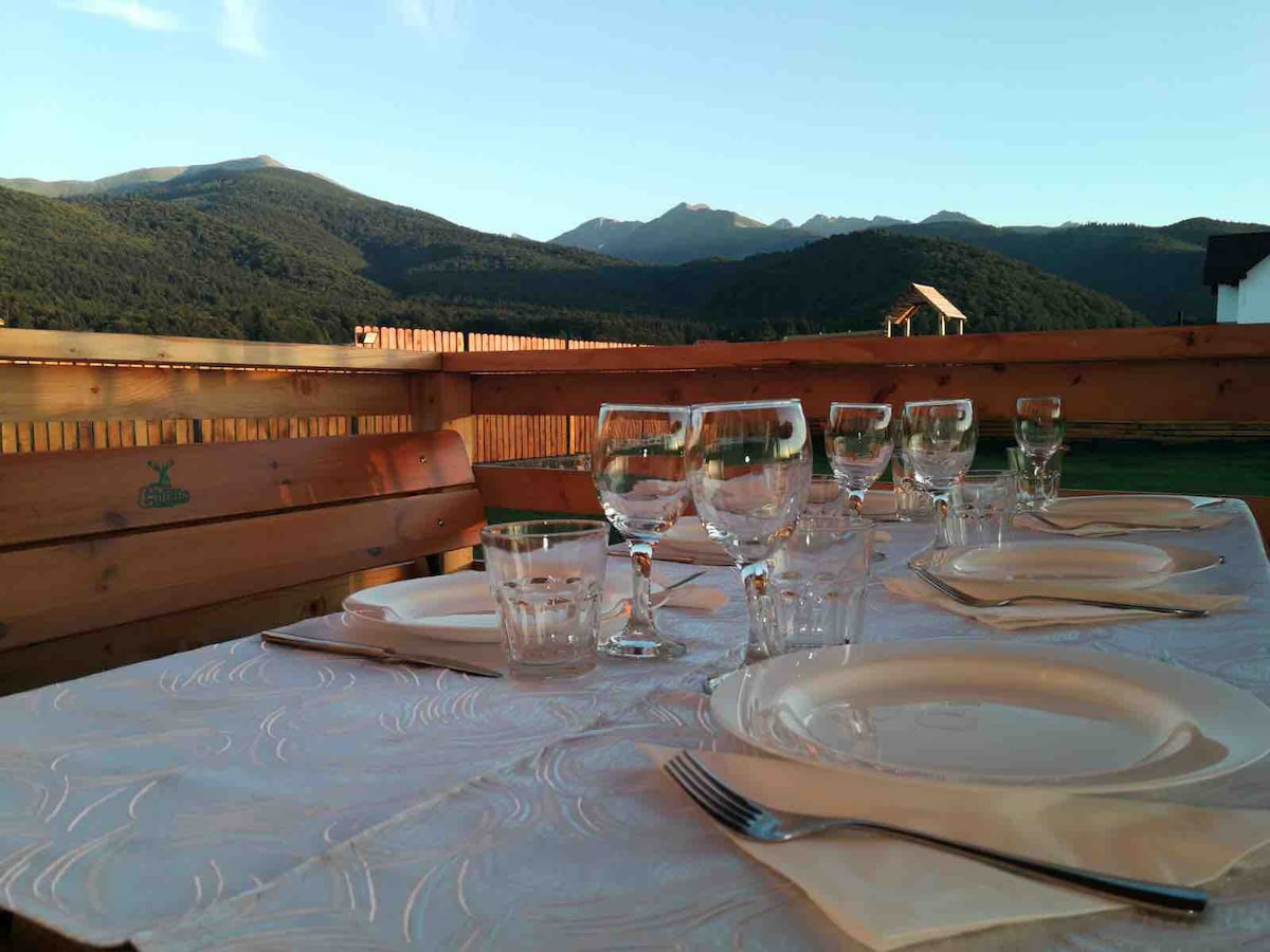 An outdoor dining table is set with glassware and plates, positioned against a backdrop of rolling mountains. The wooden surroundings create a natural ambiance, while the clear sky above contributes to a tranquil atmosphere.