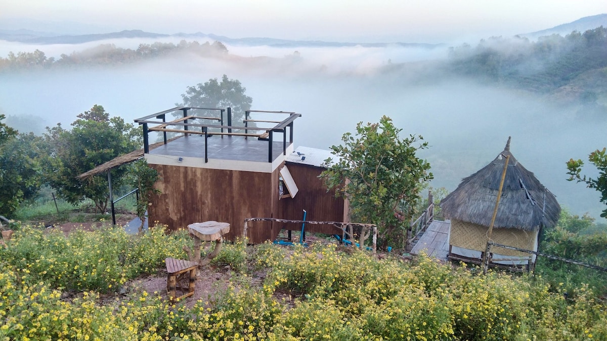 A mountain retreat is surrounded by mist, featuring a wooden structure with a rooftop deck. Nearby, a thatched-roof hut adds to the charm of the landscape. Colorful wildflowers bloom in the foreground, enhancing the serene ambiance of the natural setting.