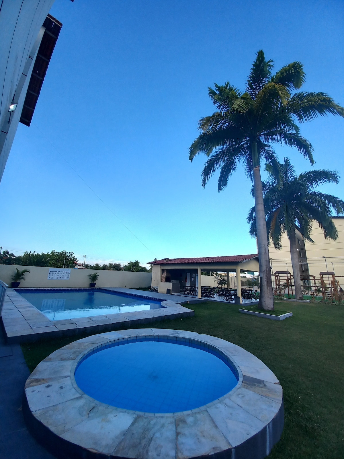 An outdoor area features two swimming pools, one larger and one smaller, both set within a landscaped yard. Tall palm trees provide shade, and a covered seating area is visible in the background, composed of a wooden structure with tables and chairs.