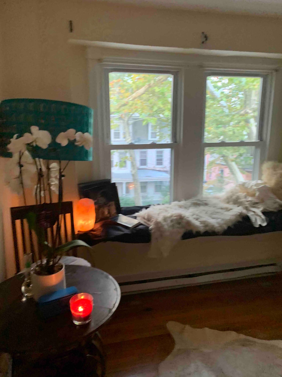 A cozy window nook features a comfortable cushion positioned beneath large windows, allowing natural light to fill the space. A small table holds a plant and a flickering candle, while soft textiles add texture. Greenery outside is visible, enhancing the tranquil environment.