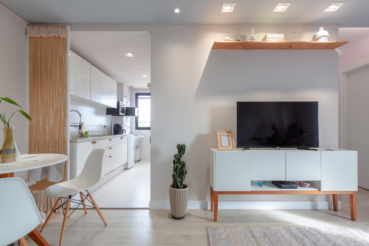 A modern living area features a white entertainment unit beneath a flat-screen TV. A small dining table with two chairs is visible adjacent to a doorway leading into a bright kitchen. Soft lighting enhances the neutral color palette, while a potted plant adds a touch of greenery.