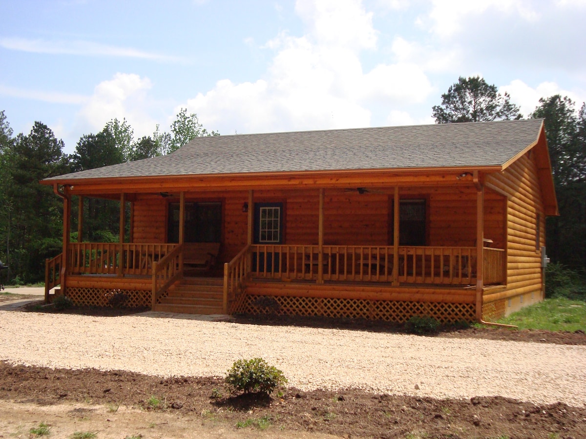 A log cabin-style house features a spacious front porch with sturdy railings and multiple seating areas. The exterior showcases natural wood tones and a shingled roof, surrounded by a gravel pathway and lush greenery. Large windows allow for natural light in the living spaces.